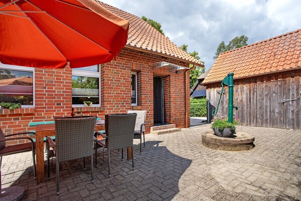 Un bâtiment en briques surmonté d’un toit en tuiles, avec des tables et des chaises en osier à l’extérieur, et un parasol rouge.