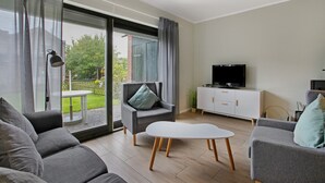 Moderni olohuone, jossa on sohva, nojatuoli ja sohvapöytä, televisio sekä liukuovet, jotka johtavat ulkotilaan.