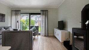 Moderni olohuone, jossa on sohva, televisio ja suuret ikkunat, joista avautuu näkymä talolle.
