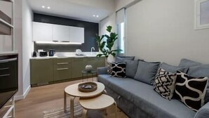 Una sala de estar moderna con un sofá gris, dos mesas redondas y una cocina al fondo.