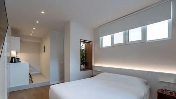 A modern bedroom with a large bed, a bedside table, a nightstand, a mirror, and a window with blinds. - Central Bliss Suite D1 (Athens)