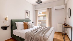 A neatly arranged bedroom with a green upholstered bed, a bedside table with a lamp, a framed picture on the wall, and a view of the outside balcony through the window. - Vibrant Living in Chalandri (Chalandri)
