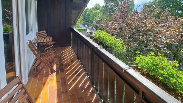 A wooden balcony with a tiled floor and a railing, overlooking a garden with various plants and trees. - Vacation apartment Schanzenblick C8 (Oberaudorf)