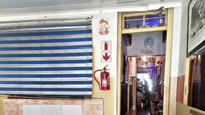 Een hotellobby met een zwart-wit geruite vloer, een zithoek met plastic stoelen, een brandblusser en een klok aan de muur.