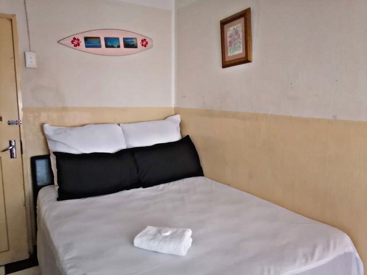 Una cama bien tendida con ropa de cama blanca y negra, una toalla blanca sobre la cama, una puerta con manija, y una pared con un cuadro y un adorno.