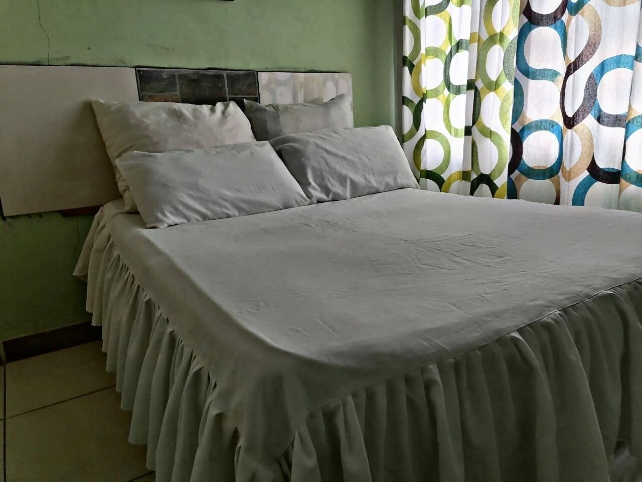 Una cama con colcha y almohadas blancas, cabecera y cortina con estampado geométrico.