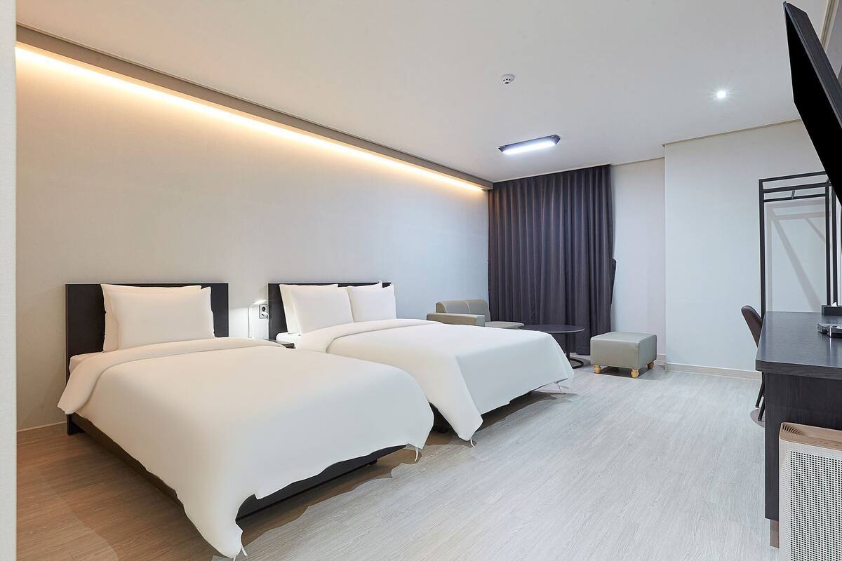 Une chambre d’hôtel avec deux lits, un bureau, une chaise, une petite table et une grande fenêtre avec des rideaux.