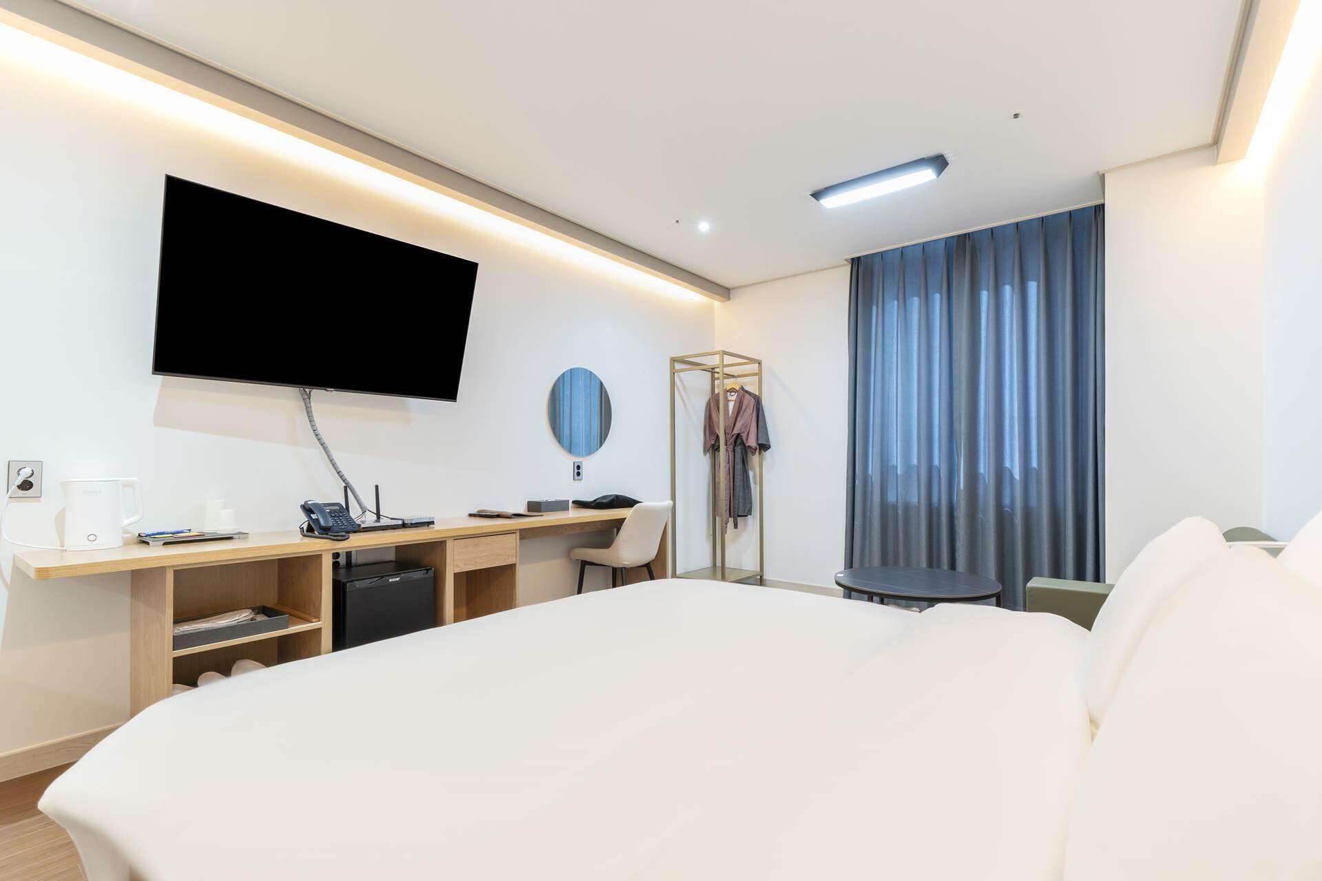 A hotel room with a bed, a desk with a computer, a flat-screen TV mounted on the wall, and a window with curtains.