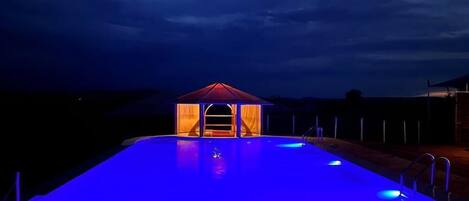 A swimming pool with a covered area and a stage, surrounded by a fence, under a dark sky.