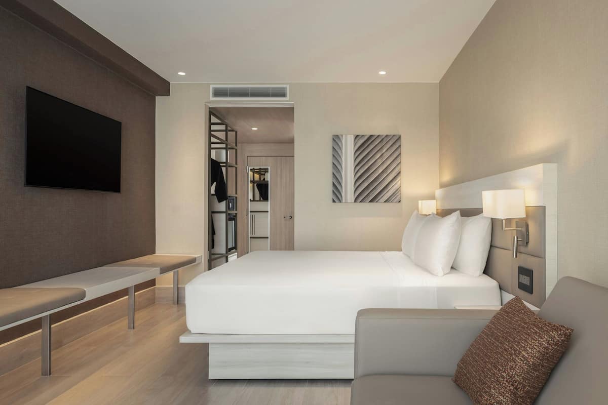 A modern hotel room with a large bed, a flat-screen TV mounted on the wall, a sofa, and a bench.