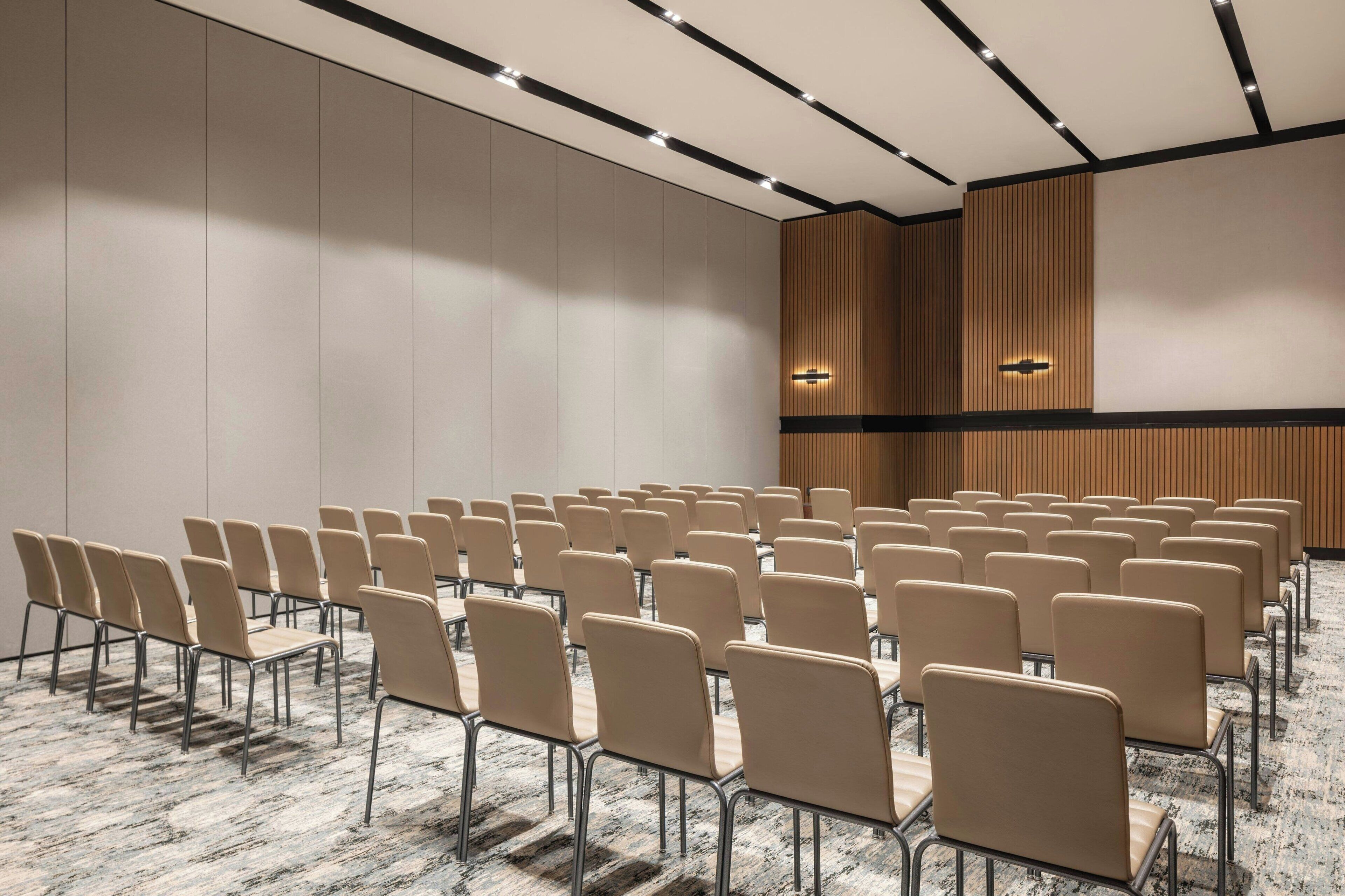 A conference room with rows of beige chairs, a wooden panel wall, and recessed ceiling lighting.