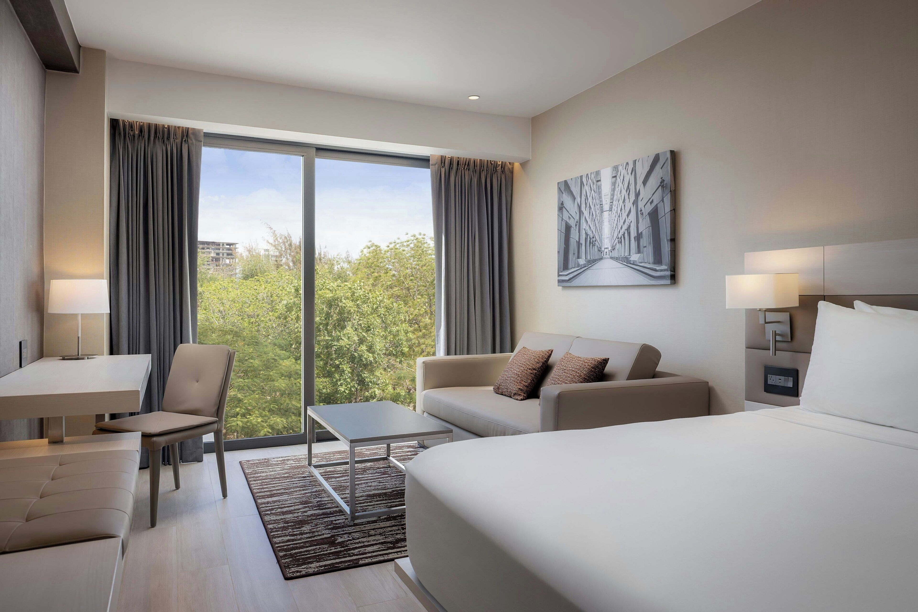 Una habitación de hotel moderna con una cama grande, un sofá, un escritorio y una silla. Cuenta con un ventanal con cortinas, un cuadro en la pared y una mesita de noche con lámpara.
