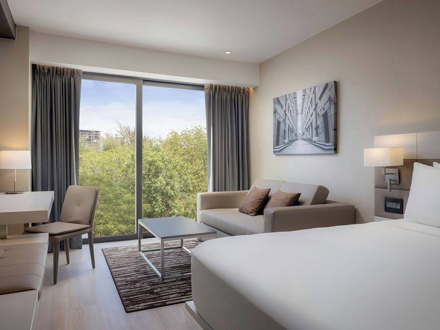 A modern hotel room with a large bed, a sofa, a desk, and a chair. There is a large window with curtains, a painting on the wall, and a nightstand with a lamp.