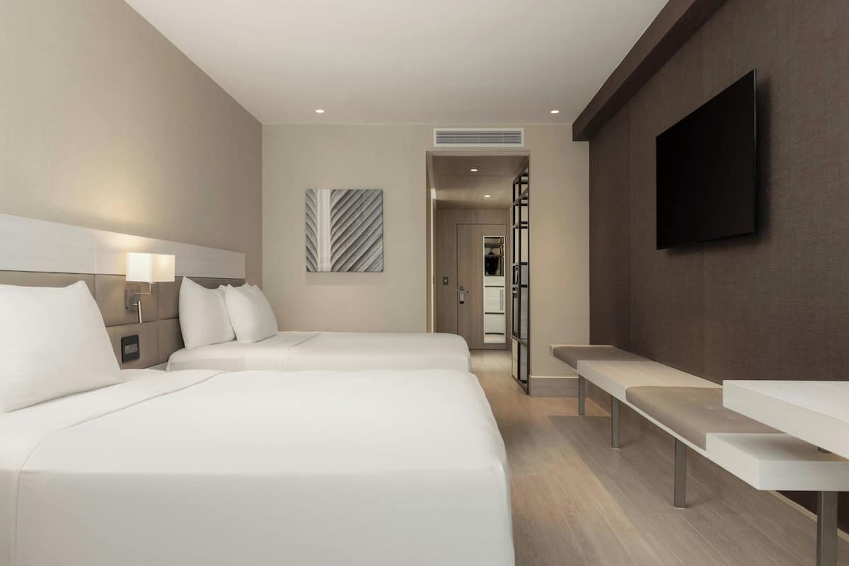 A hotel room with two beds, a TV mounted on the wall, and a bench.