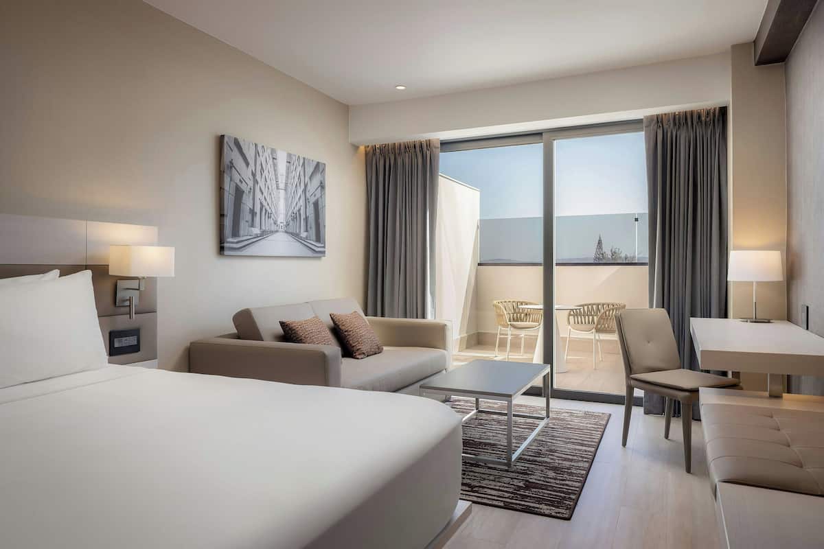 A modern hotel room with a large bed, a sofa, a dining table, and a balcony with a view.