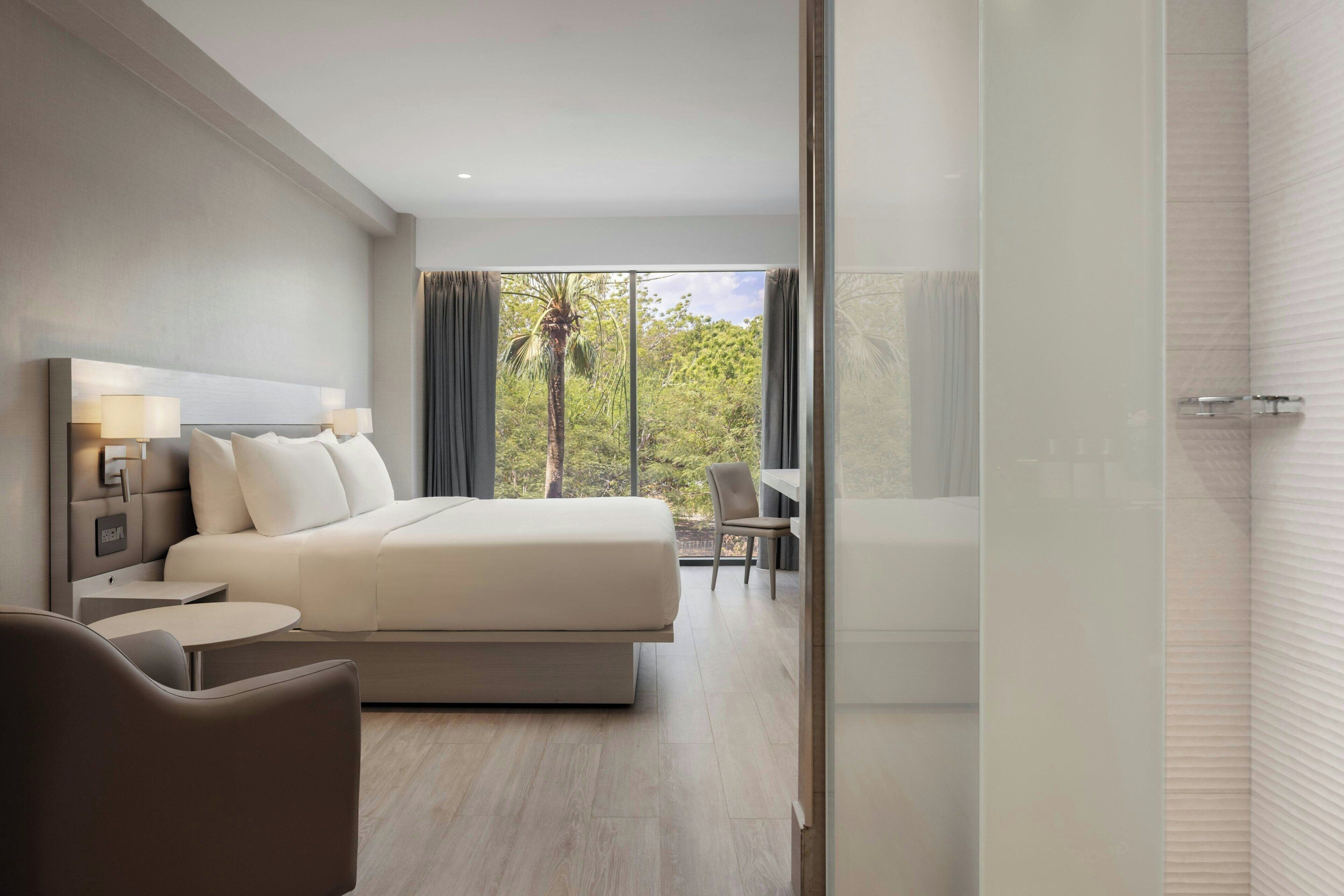 Habitación de hotel moderna con una cama grande, mesitas de noche, una silla y vista a zonas verdes por la ventana.