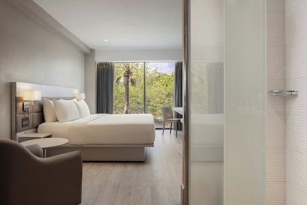 Habitación de hotel moderna con una cama grande, mesitas de noche, una silla y vista a zonas verdes por la ventana.