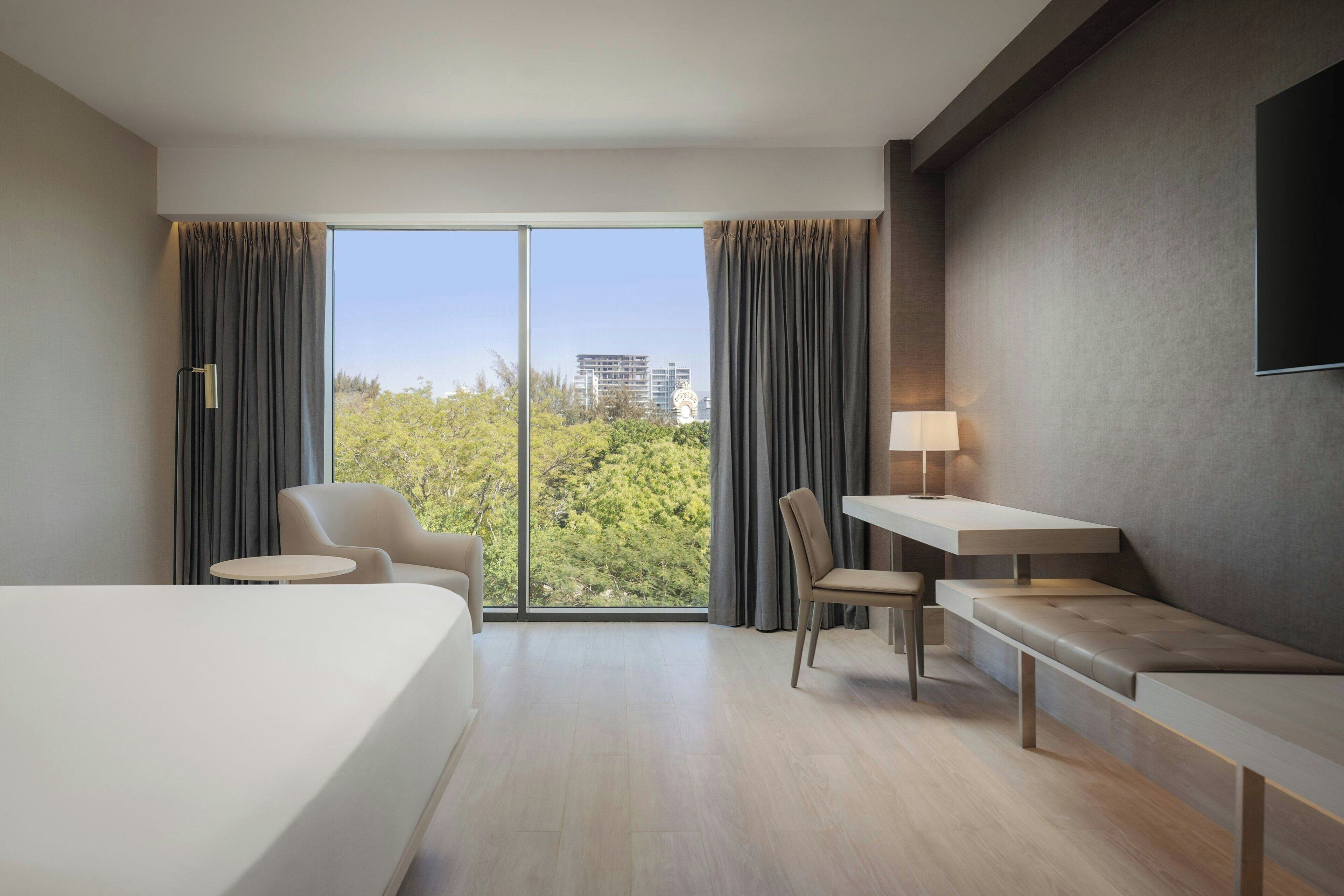 Ein modernes Hotelzimmer mit einem großen Bett, einem Schreibtisch, einem Sessel und einer Couch. Ein Fenster mit Vorhängen, eine Lampe und ein an der Wand befestigter Fernseher sind vorhanden.