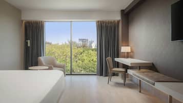 Una habitación de hotel moderna con una cama grande, un escritorio, una silla y un sofá. Hay una ventana con cortinas, una lámpara y un televisor montado en la pared.