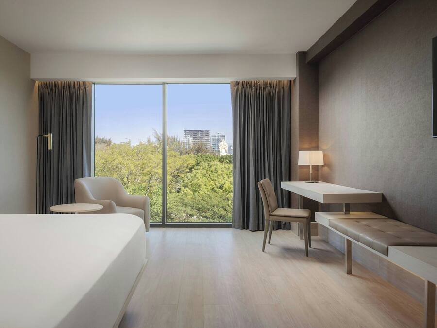 A modern hotel room with a large bed, a desk, a chair, and a sofa. There is a window with curtains, a lamp, and a TV mounted on the wall.