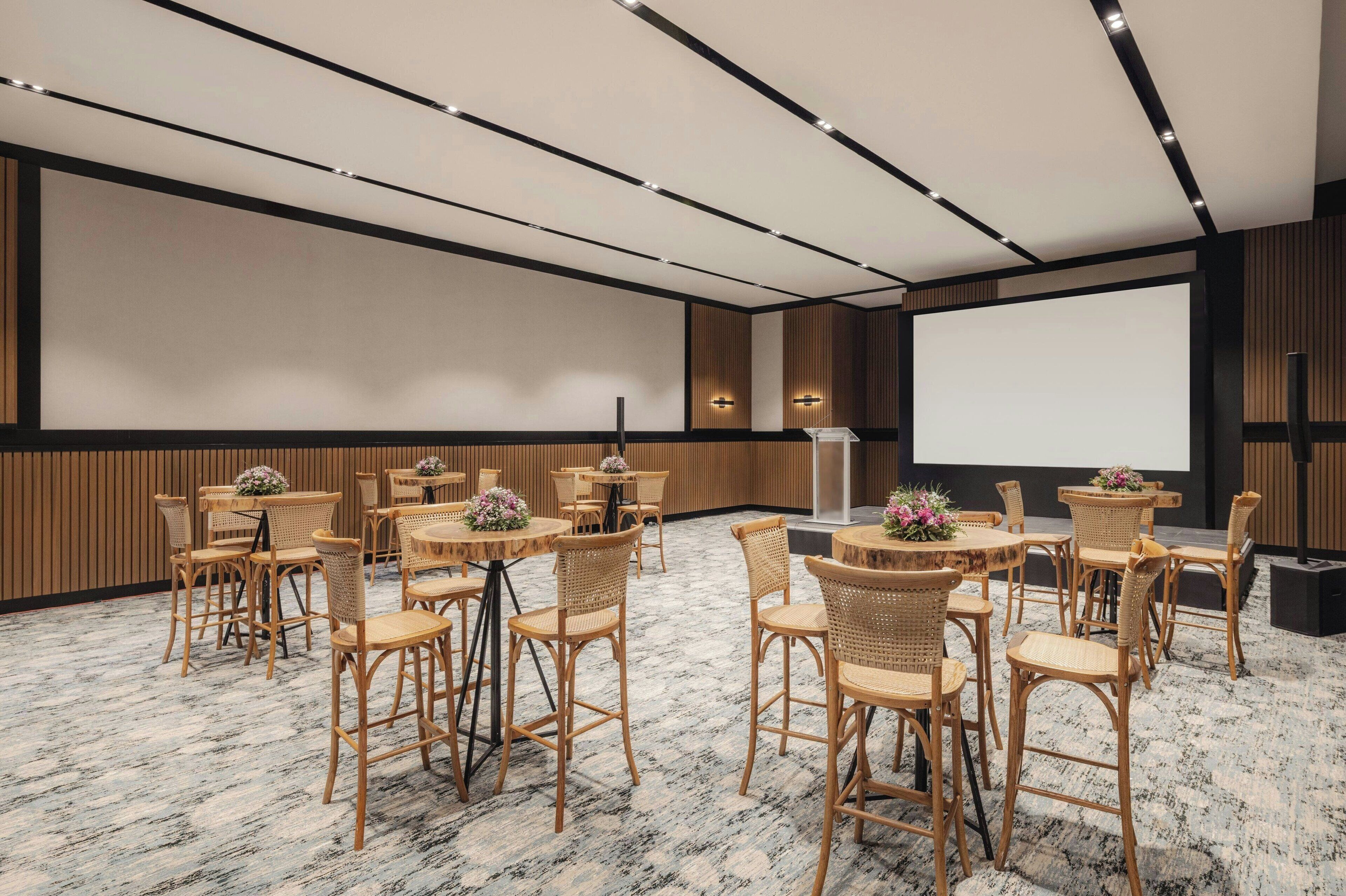 Un salón de conferencias con varias mesas redondas y sillas de mimbre, equipado con una pantalla de proyector e iluminación empotrada.