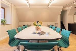 Un salón de conferencias con una mesa blanca, sillas color turquesa, un libro, un jarrón con flores y una taza de café.