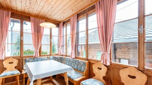 A wooden interior with a table and benches, large windows with curtains, and a view of the outside.
