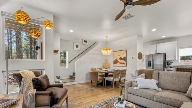 A spacious living room with a sofa, a coffee table, and a dining area with chairs and a table. There is a staircase leading to another level, and a kitchen area with cabinets and a refrigerator.