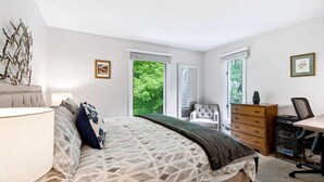 Ein Schlafzimmer mit Bett, Kommode, Schreibtisch und Stuhl. Es gibt Fenster mit Jalousien, eine Lampe und ein dekoratives Wandbild.