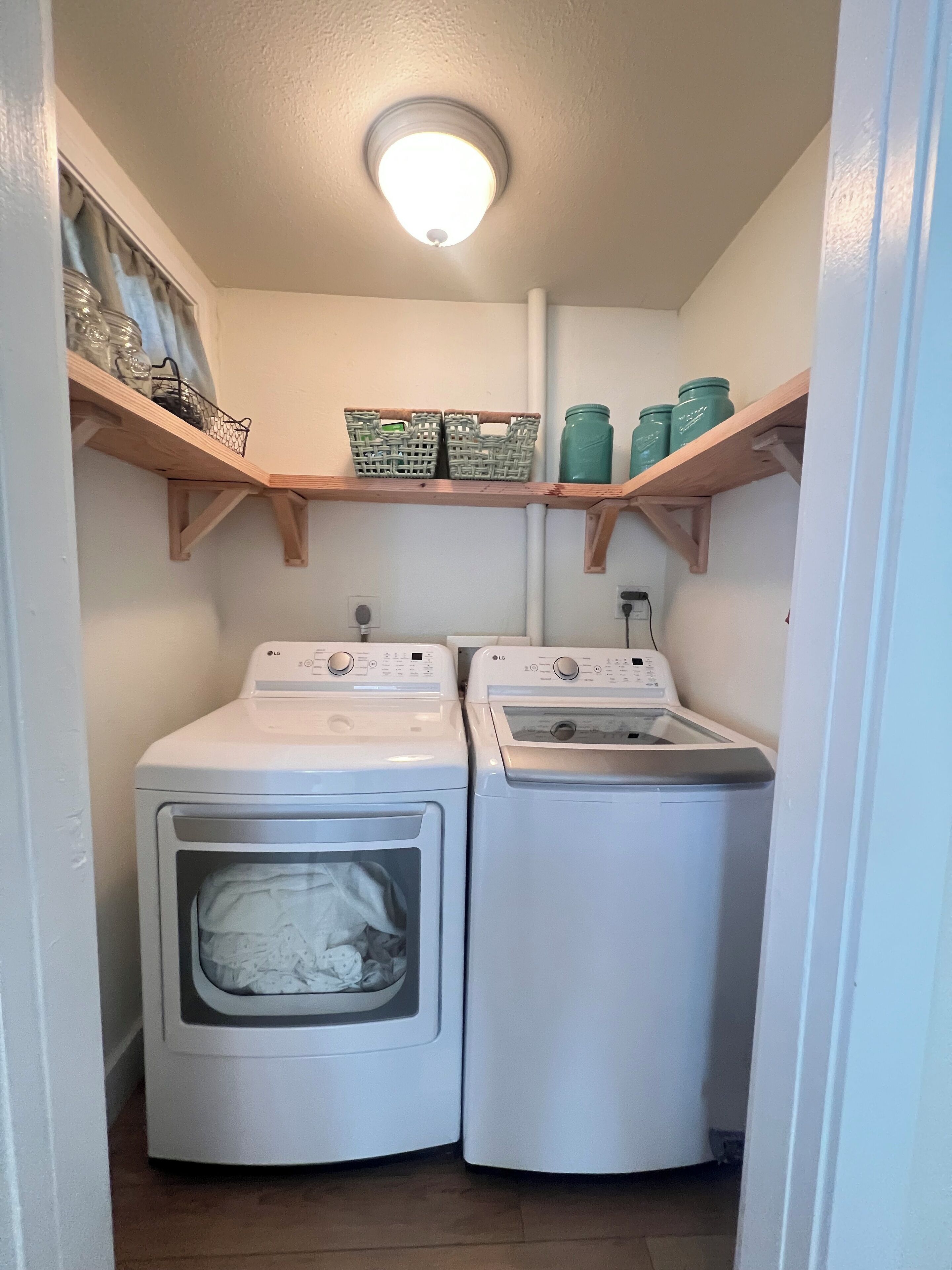 A laundry room with two front-loading washing machines, wooden shelves above holding baskets and jars, and a ceiling light.