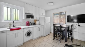 A modern kitchen with white cabinets, a washing machine, a microwave, and a refrigerator. There is a dining table with chairs and a window with blinds. - Lovely ground floor suite in Central Vancouver (Vancouver)