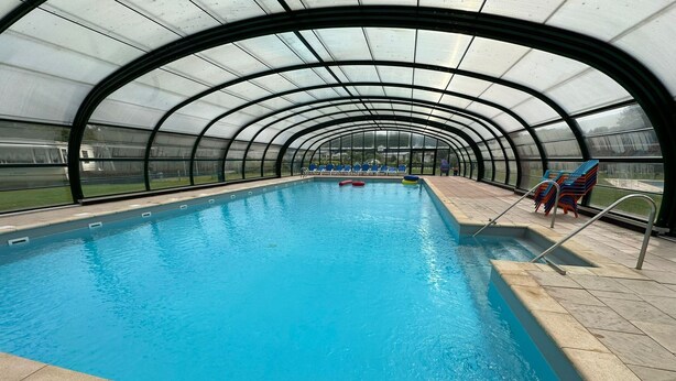 An indoor swimming pool with a curved roof and clear glass walls, surrounded by blue chairs and a pool ladder. - Holiday home for group with sauna and pool (Havelte)