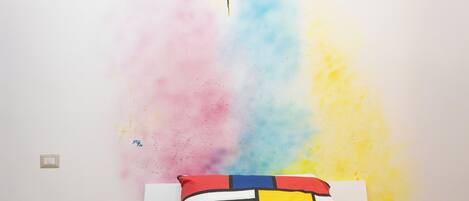A bed with a colorful geometric-patterned bedspread and pillows, a white wall with a colorful abstract design, and two bedside tables with decorative items.