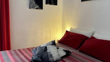 Un dormitorio con una colcha a rayas, un almohón rojo y una pared decorada con cuatro cuadros.