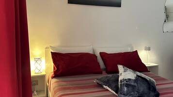 Un lit avec une literie à rayures, des oreillers rouges et un oreiller noir et blanc. Il y a une table de chevet avec une lampe et une applique murale. Un tableau est accroché au-dessus du lit.