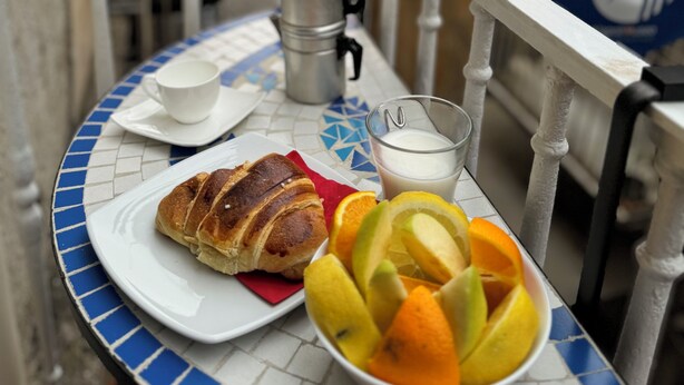 A table with a croissant, a glass of milk, and a bowl of fruit on a tiled table in an outdoor setting. - Napoli Museo Bed and Breakfast (Naples)