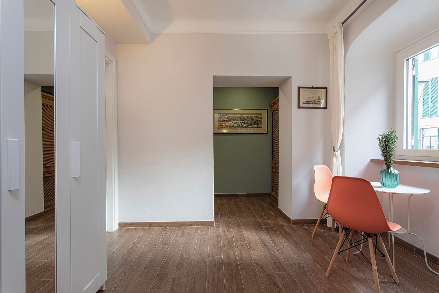 A hallway with wooden flooring, a white door, a framed picture on the wall, and a window with a view of buildings.