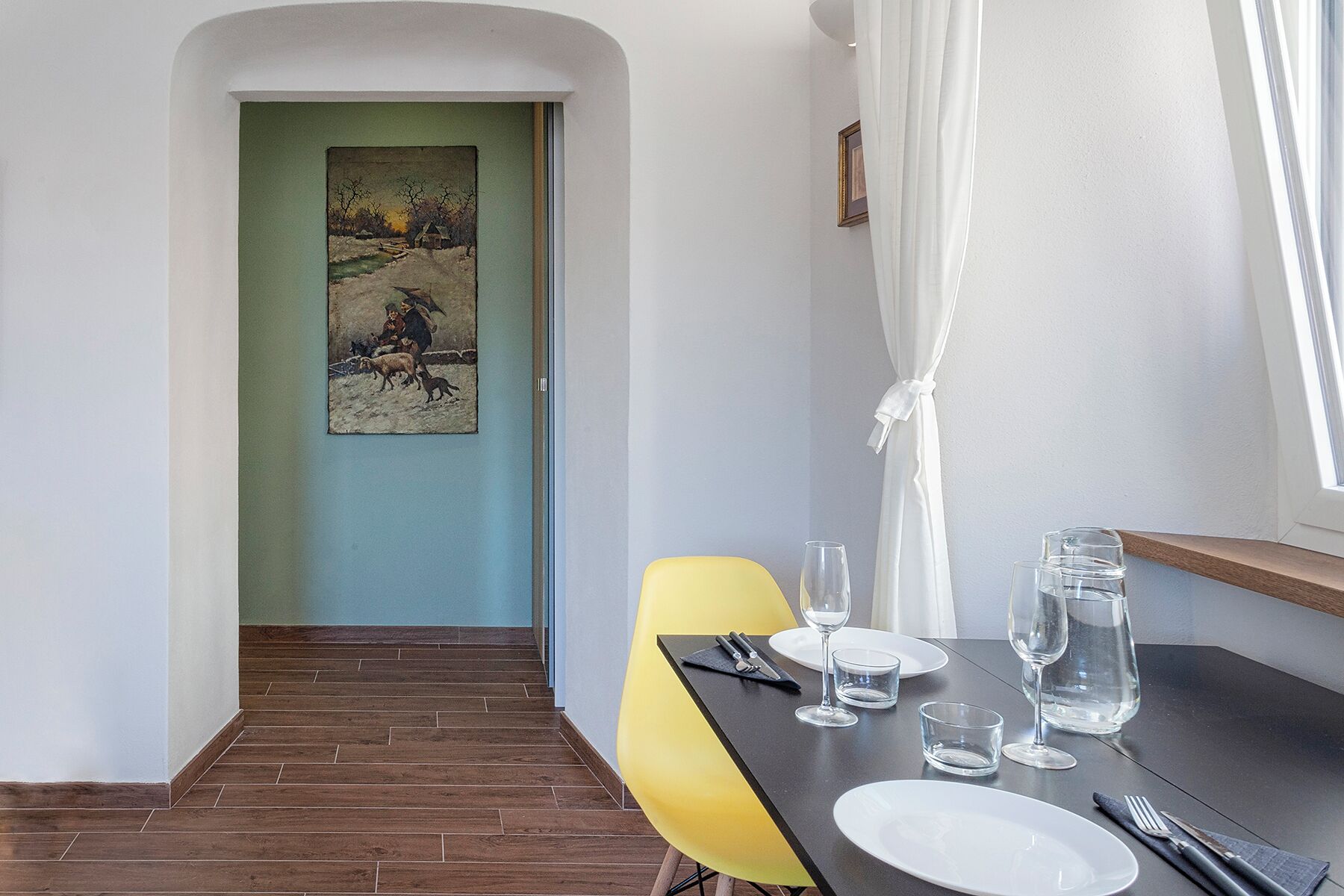 A dining area with a wooden floor, a yellow chair, a black table set for two, and a painting of a winter scene on the wall.