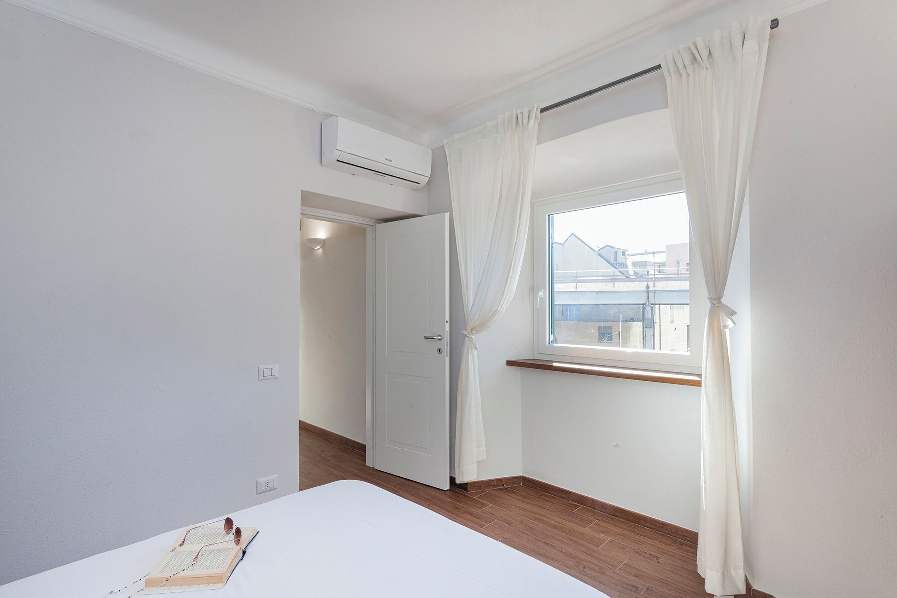 A bedroom with a wooden floor, a white bed, a window with white curtains, and an air conditioning unit on the wall.