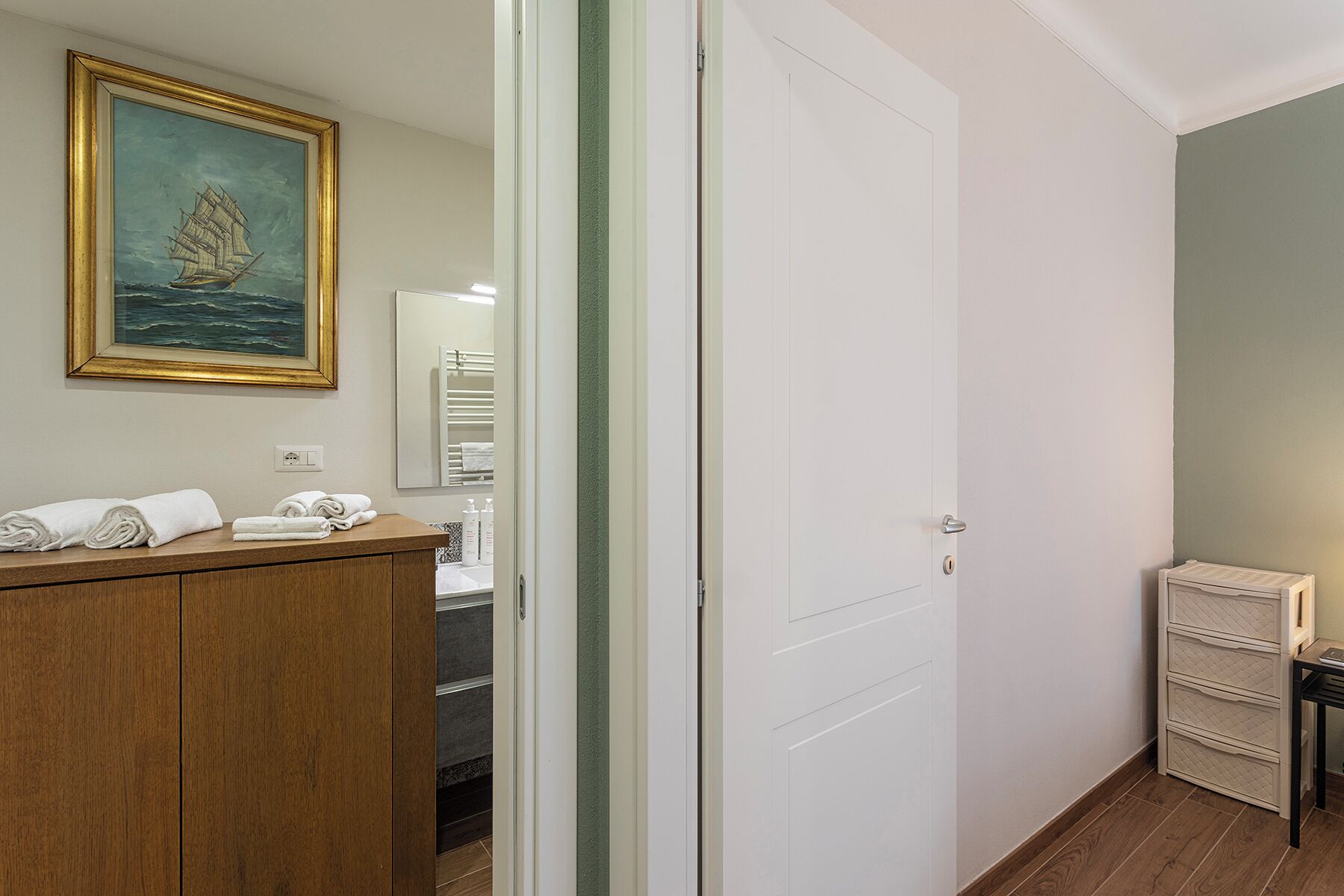 Ein Hotelzimmer mit einem Holznächtigstisch, einem gerahmten Gemälde eines Schiffes, einer weißen Tür und einem kleinen Schrank mit Schubladen.