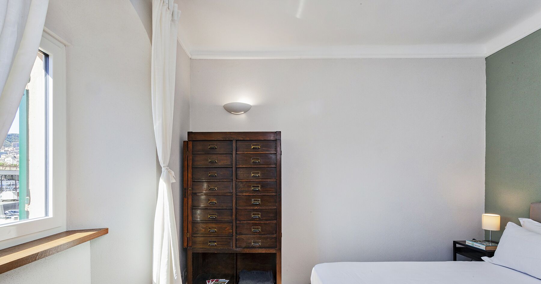 A bedroom with a bed, a nightstand with a lamp, a wooden dresser with multiple drawers, a window with curtains, and a view of a cityscape.