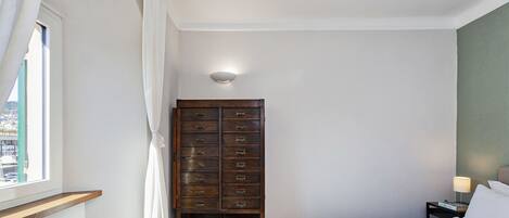 Un dormitorio con cama, una mesita de noche con lámpara, un aparador de madera con varios cajones, una ventana con cortinas y vista a la ciudad.