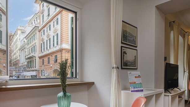 A hotel room with a large window offering a view of historic buildings, a small table with a vase, and a desk with a computer and a chair. - San Lorenzo View by Wonderful Italy (Genoa)