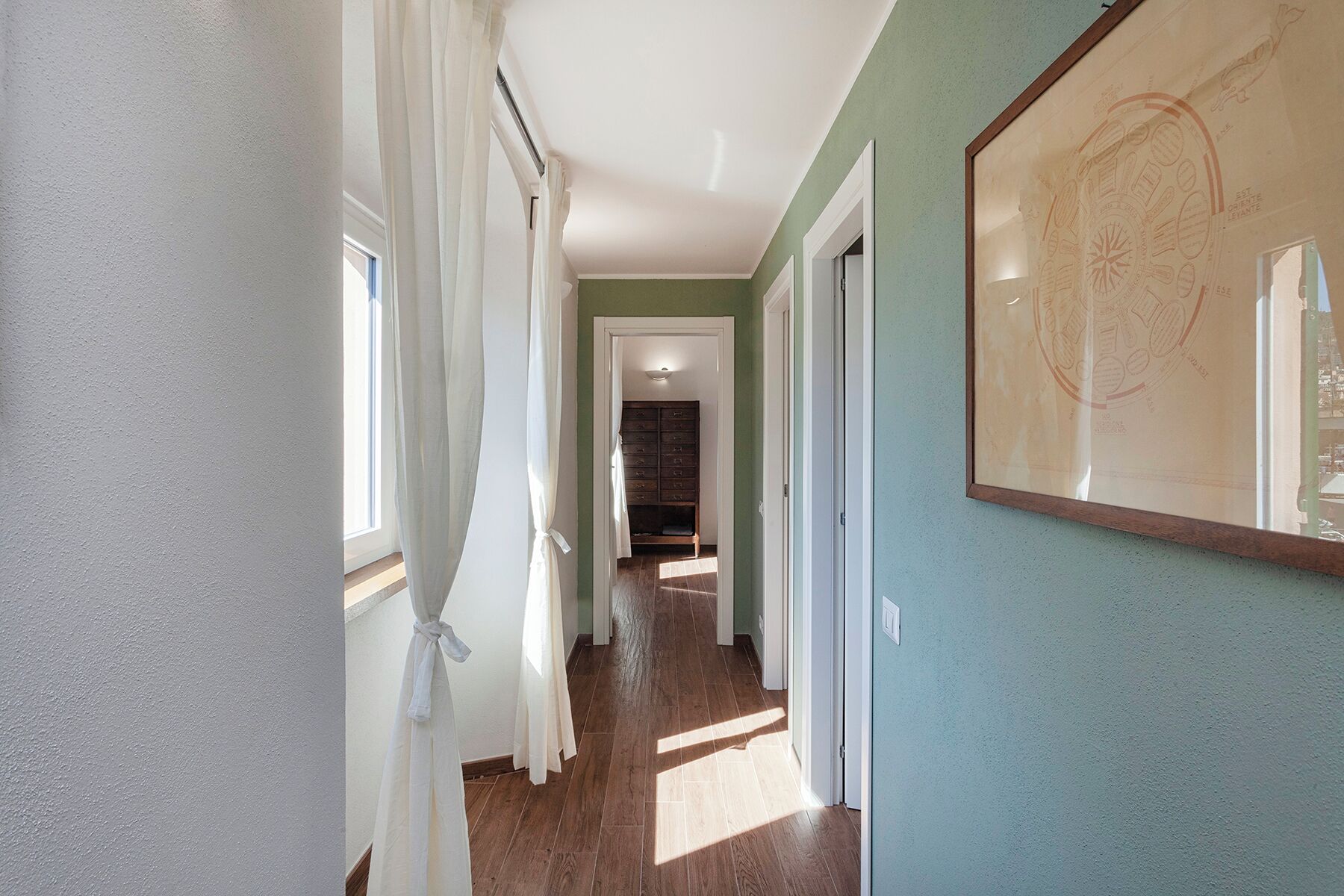 A hallway with wooden flooring, white curtains, and a framed map on the wall.