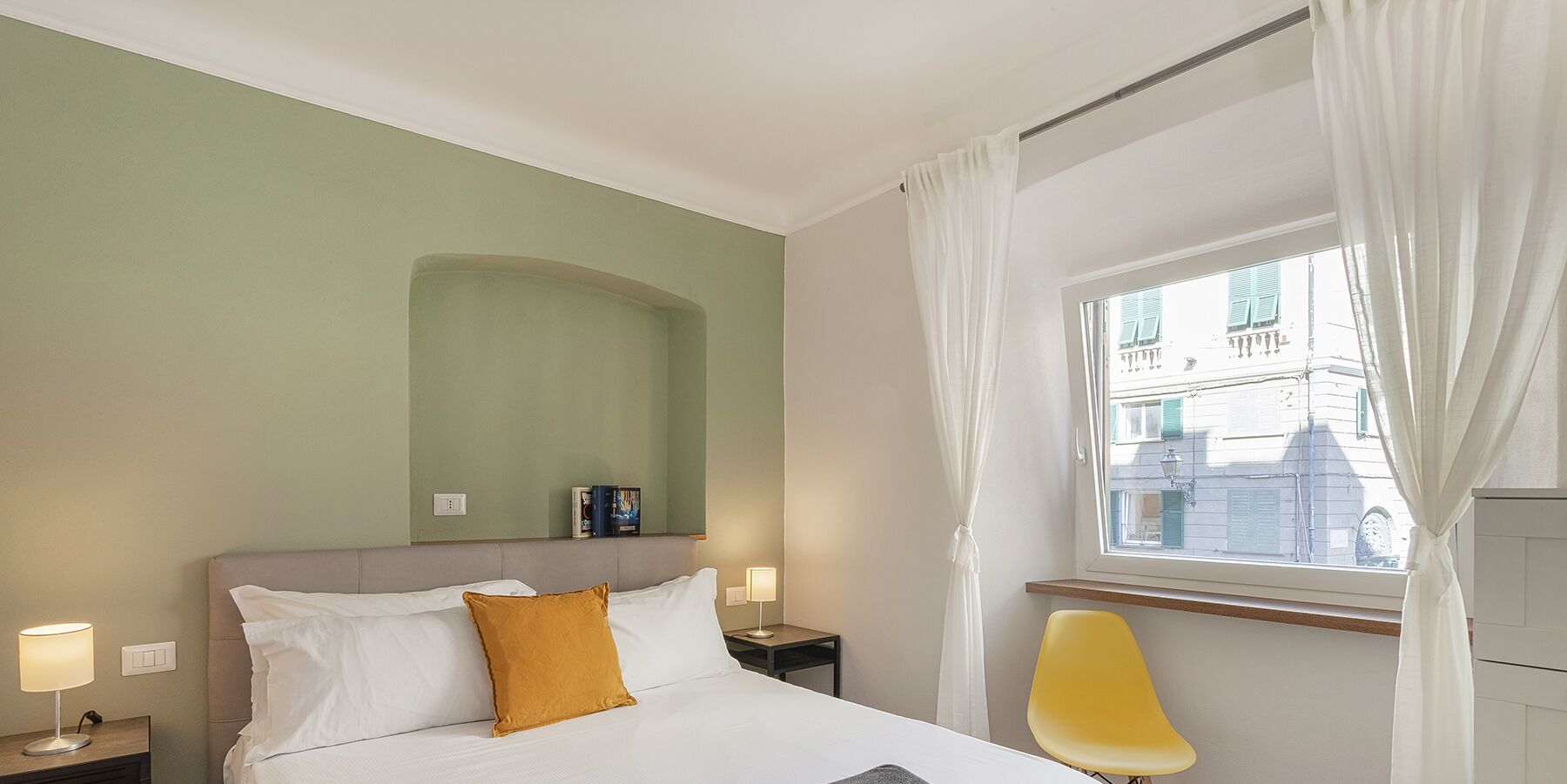 A hotel room with a bed, a nightstand, a lamp, a chair, a window with curtains, and a view of buildings outside.