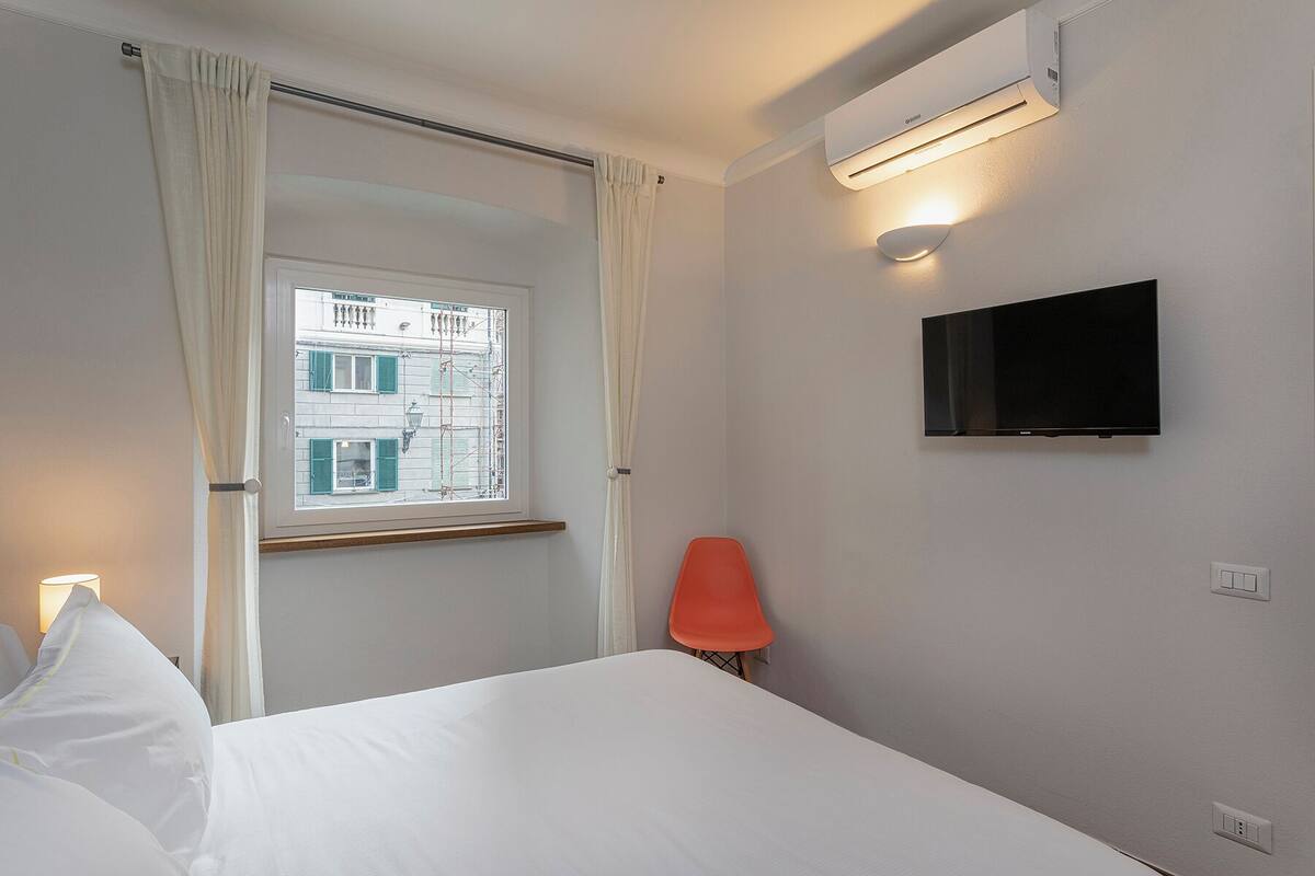 A hotel room with a bed, a TV mounted on the wall, a window with curtains, and an air conditioning unit.
