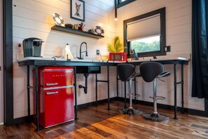 Une cuisine compacte avec un réfrigérateur rouge, un four à micro-ondes et un évier. On y trouve deux tabourets de bar noirs et une fenêtre donnant sur un paysage verdoyant.