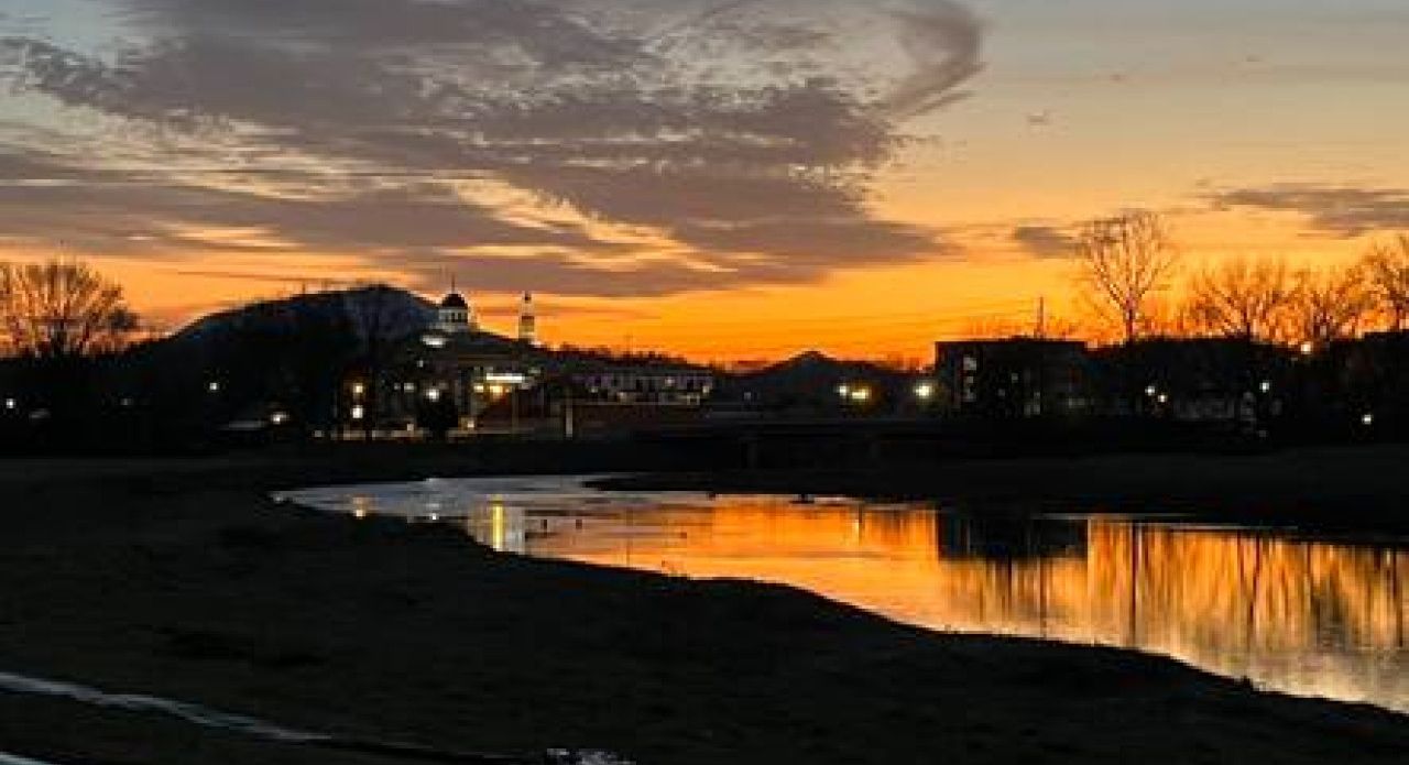 Ein Sonnenuntergang über einem Fluss, am gegenüberliegenden Ufer stehen Gebäude.