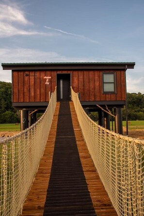 Un pont suspendu en bois mène à un chalet doté d’un toit sombre et d’une petite fenêtre.