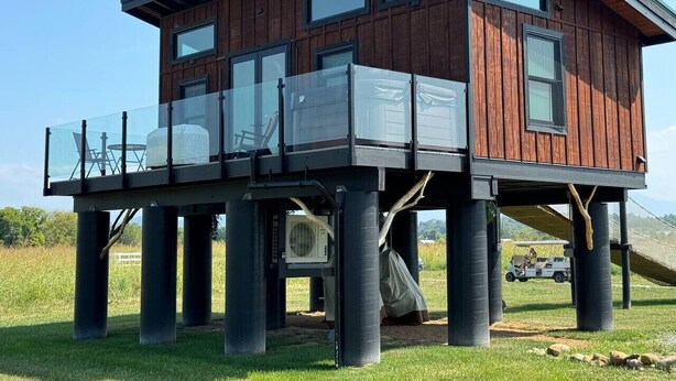 Une maison en bois dotée d’un balcon, soutenu par des piliers, et un véhicule visible à l’arrière-plan.